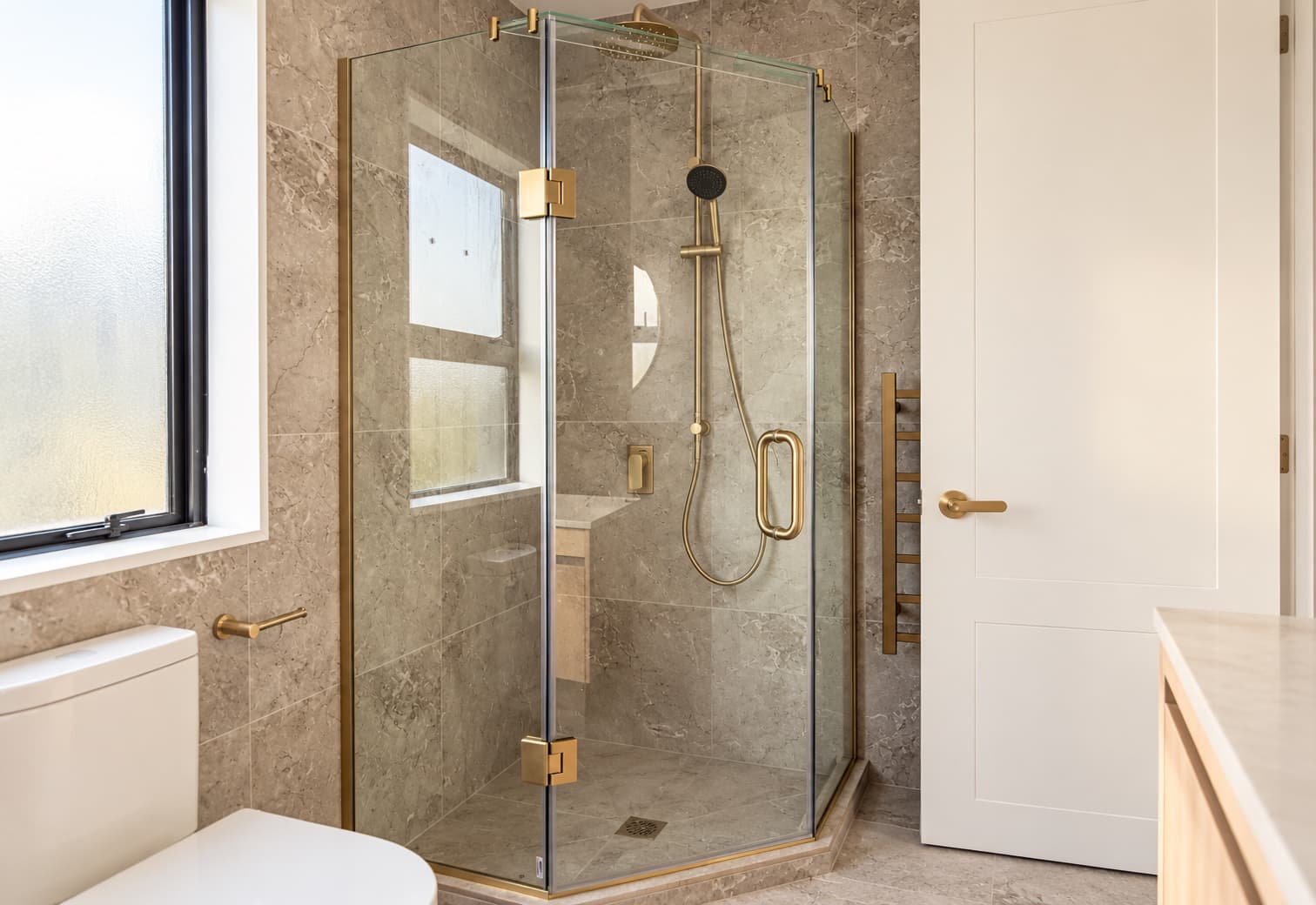 Frameless glass shower box with gold hardware in a stone-tiled bathroom
